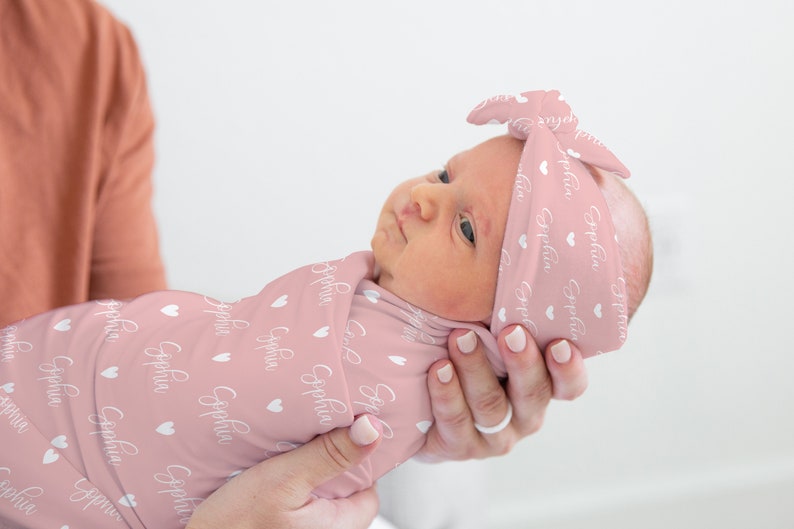 May include: A pink swaddle blanket with white hearts and the name "Sophia" printed on it. A baby is wrapped in the blanket and wearing a matching pink headband with the same pattern.