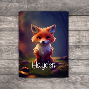 May include: A rectangular blanket with a cartoon fox in orange and white fur, sitting on grass. The blanket has the name "Hayden" in white script. The background is a blurred forest scene.