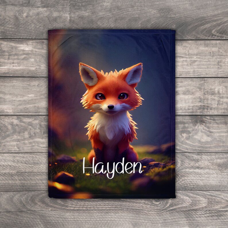 May include: A rectangular blanket with a cartoon fox in orange and white fur, sitting on grass. The blanket has the name "Hayden" in white script. The background is a blurred forest scene.