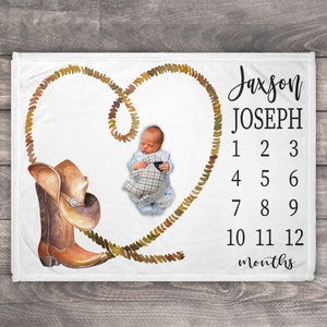 May include: A white baby blanket with a brown cowboy boot and a lasso forming a heart shape. The blanket has the name "Jaxson Joseph" and numbers 1-12 for tracking a baby's growth.