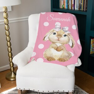 Op de afbeelding: Een roze deken met witte stippen en een bruin konijn dat een roze strik draagt. De deken is gepersonaliseerd met de naam "Savannah".