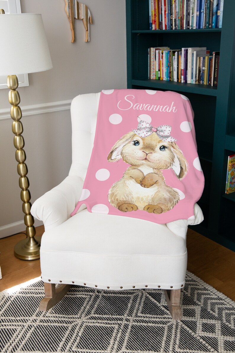 Op de afbeelding: Een roze deken met witte stippen en een bruin konijn dat een roze strik draagt. De deken is gepersonaliseerd met de naam "Savannah".
