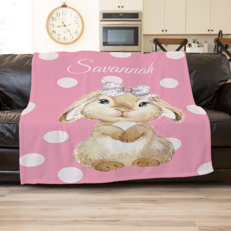 Op de afbeelding: Een roze fleece deken met witte stippen en een cartoon konijn met een roze strik. De deken heeft de naam "Savannah" erop gedrukt.