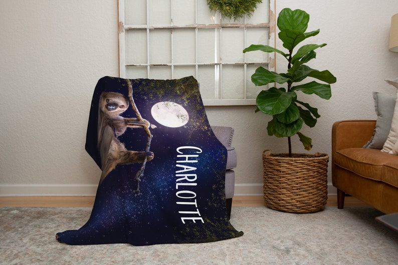 May include: A soft, blue fleece blanket featuring a sloth hanging from a branch in front of a full moon. The blanket has a starry night background and the name "Charlotte" printed on the side.