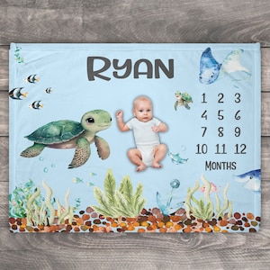 May include: A blue baby milestone blanket with a sea turtle, fish, and an anchor. The blanket has a personalized name "RYAN" and numbers 1-12 for each month.