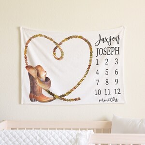 May include: White baby milestone blanket with a brown cowboy boot and hat design. The blanket has a rope heart design and the text "Jaxson Joseph 1 2 3 4 5 6 7 8 9 10 11 12 months".