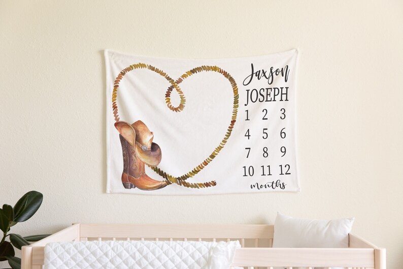 May include: White baby milestone blanket with a brown cowboy boot and hat design. The blanket has a rope heart design and the text "Jaxson Joseph 1 2 3 4 5 6 7 8 9 10 11 12 months".