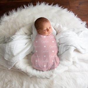 May include: A pink swaddle blanket with white hearts and the name "Sophia" printed on it. A baby is swaddled in the blanket and is lying on a white fluffy surface.