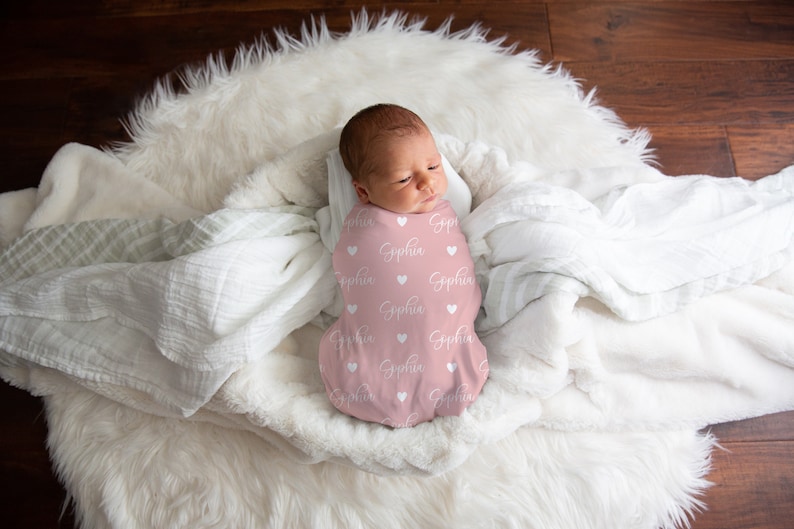 May include: A pink swaddle blanket with white hearts and the name "Sophia" printed on it. A baby is swaddled in the blanket and is lying on a white fluffy surface.