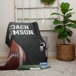 Pu&ograve; includere: Una coperta in pile nera e grigia con il testo "COACH SAMSON" in bianco. Un pallone da calcio e un fischietto sono anche raffigurati sulla coperta.