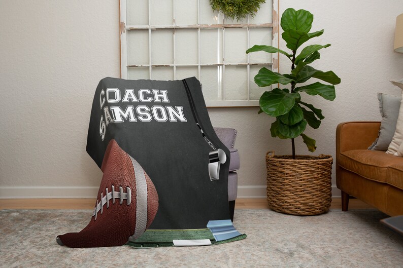 Pu&ograve; includere: Una coperta in pile nera e grigia con il testo "COACH SAMSON" in bianco. Un pallone da calcio e un fischietto sono anche raffigurati sulla coperta.