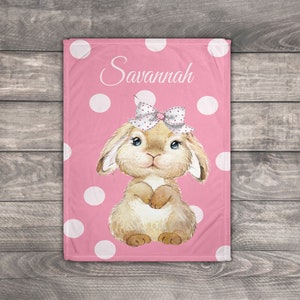 Op de afbeelding: Een roze babydeken met witte stippen en een bruin konijn met een roze strik en de naam "Savannah" erop.