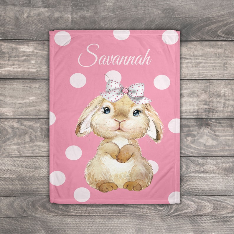 Op de afbeelding: Een roze babydeken met witte stippen en een bruin konijn met een roze strik en de naam "Savannah" erop.