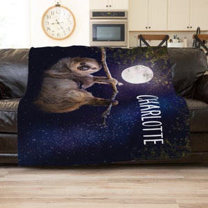 May include: A blue and purple fleece blanket featuring a sloth hanging from a branch in front of a full moon. The blanket has a starry night sky background and the name "CHARLOTTE" is printed in white on the bottom right corner.
