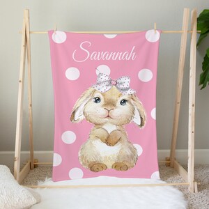 Op de afbeelding: Een roze deken met witte stippen en een bruin konijn met een roze strik en de naam "Savannah" erop.
