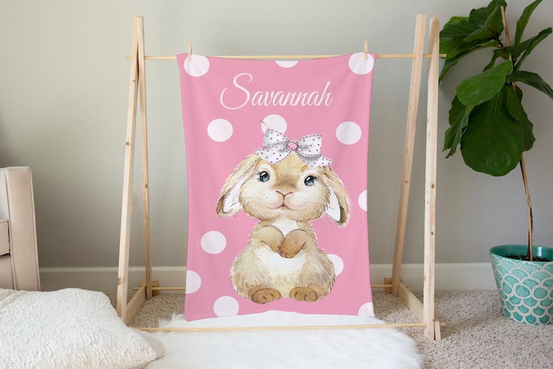 Op de afbeelding: Een roze deken met witte stippen en een bruin konijn met een roze strik en de naam "Savannah" erop.