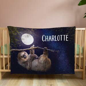 May include: A blue and black personalised baby blanket featuring a sloth hanging from a branch beneath a full moon. The blanket has a starry night sky background and the name "CHARLOTTE" is printed in white.