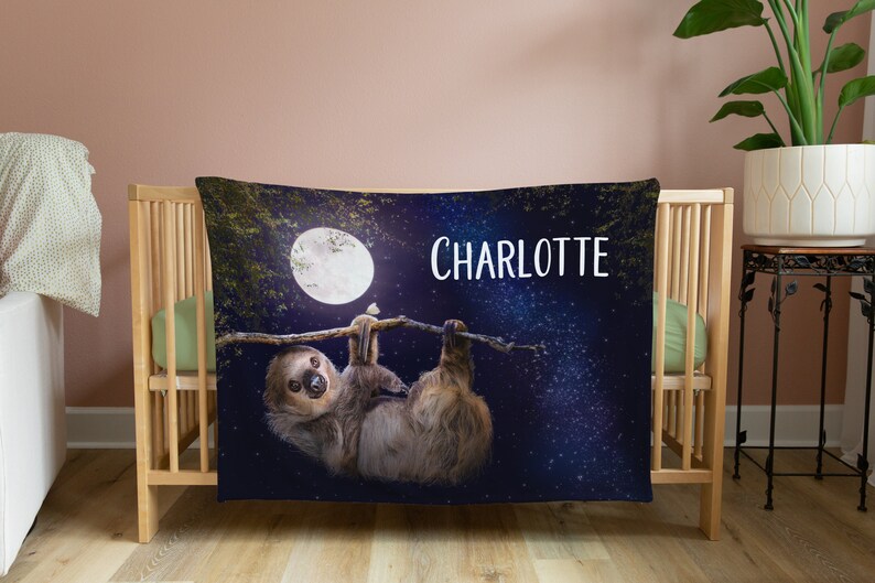 May include: A blue and black personalised baby blanket featuring a sloth hanging from a branch beneath a full moon. The blanket has a starry night sky background and the name "CHARLOTTE" is printed in white.