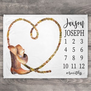 May include: A white baby blanket with a brown cowboy boot and hat, a brown rope in the shape of a heart, and the text "Jaxson JOSEPH 1 2 3 4 5 6 7 8 9 10 11 12 months".