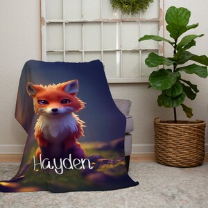 May include: A soft blanket with a cartoon fox design in orange and white, set in a forest scene. The blanket displays the name "Hayden" in white script. The blanket is draped over a chair.