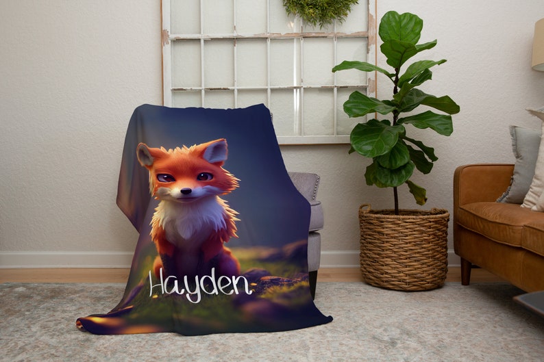 May include: A soft blanket with a cartoon fox design in orange and white, set in a forest scene. The blanket displays the name "Hayden" in white script. The blanket is draped over a chair.