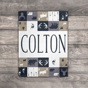 May include: A white and gray baby blanket with a patchwork design featuring woodland animals, antlers, arrows, and feathers. The name "COLTON" is printed in black on the blanket.