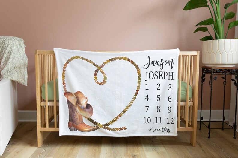 May include: White baby blanket with a brown cowboy boot and hat design. A rope is shaped into a heart with the text "Jaxson Joseph" and numbers 1-12 for each month.