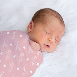 May include: A pink swaddle blanket with white hearts and the name "Sophia" printed on it. A baby is sleeping inside the swaddle.
