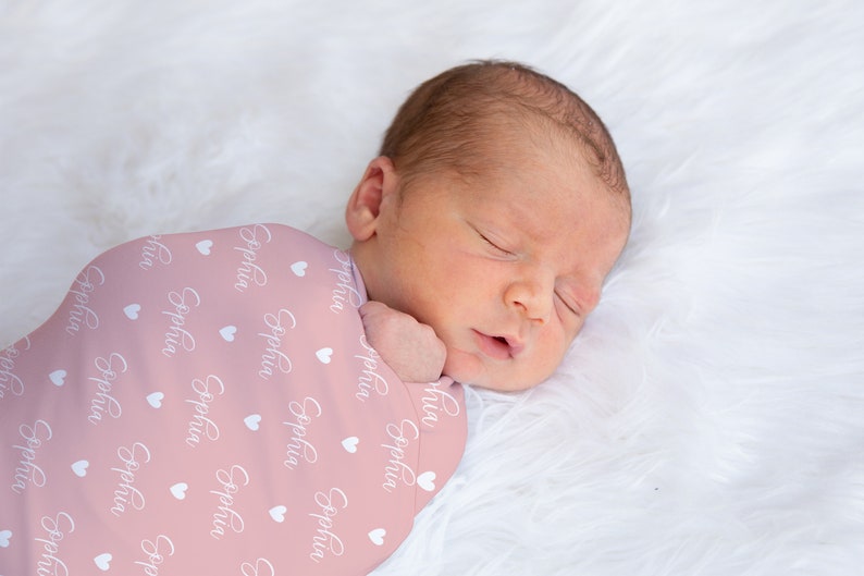 May include: A pink swaddle blanket with white hearts and the name "Sophia" printed on it. A baby is sleeping inside the swaddle.