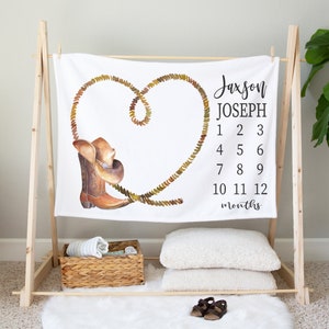 May include: White baby blanket with a brown cowboy boot and hat design. The blanket has a rope heart design and the text "Jaxson Joseph 1 2 3 4 5 6 7 8 9 10 11 12 months".