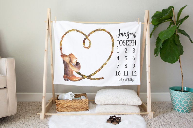 May include: White baby blanket with a brown cowboy boot and hat design. The blanket has a rope heart design and the text "Jaxson Joseph 1 2 3 4 5 6 7 8 9 10 11 12 months".