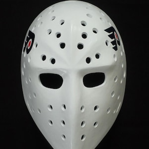 Op de afbeelding: Een witte hockey-doelmasker met talrijke ventilatiegaten. Het masker toont het teamlogo in zwart en rood aan beide zijden. Het masker heeft een gladde, glanzende afwerking en is ontworpen om het gezicht te beschermen.