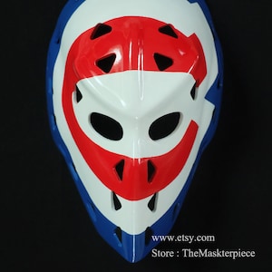 K&ouml;nnte beinhalten: Eine wei&szlig;e Hockey-Torwartmaske mit rotem und blauem Design. Die Maske hat ein gro&szlig;es "C" auf der Vorderseite, mit einem roten Streifen oben und einem blauen Streifen unten. Die Maske hat eine Reihe von L&ouml;chern zur Bel&uuml;ftung.
