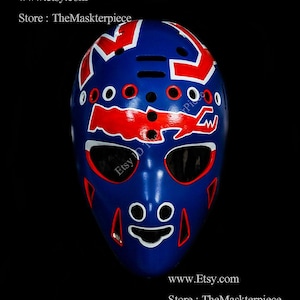 May include: A blue hockey goalie mask with red and white accents. The mask features a stylized alligator design with a white outline. The mask has a black background and a white text overlay that reads "Etsy D TheMaskterpiece".