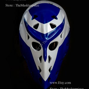 May include: A white and blue goalie mask with a unique design. The mask has a white V-shaped design with blue accents. The mask has a variety of holes for ventilation. The text "Etsy ID TheMaskterpiece" is visible on the mask. The text "www.etsy.com Store: TheMaskterpiece" is visible at the bottom of the image.