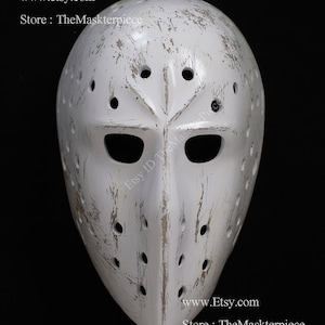 Könnte beinhalten: Weißer Hockey-Torwarthelm mit abgenutzter Lackierung. Der Helm hat ein traditionelles Design mit einer großen Augenöffnung und einer kleinen Mundöffnung. Der Helm ist mit kleinen Löchern bedeckt und hat einen abgenutzten Look.