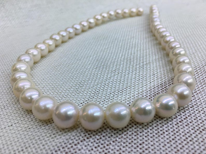 May include: A strand of lustrous, round, white pearls. The pearls are strung together, forming a curved shape. The pearls have a smooth, reflective surface, and are displayed on a textured, off-white background. The pearls are a classic jewelry item.