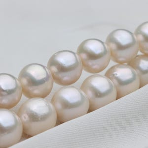May include: Close-up of a strand of lustrous, creamy white pearls. The pearls are round and evenly sized, with a subtle iridescence. The pearls are arranged diagonally against a white fabric background, suggesting a necklace or bracelet.