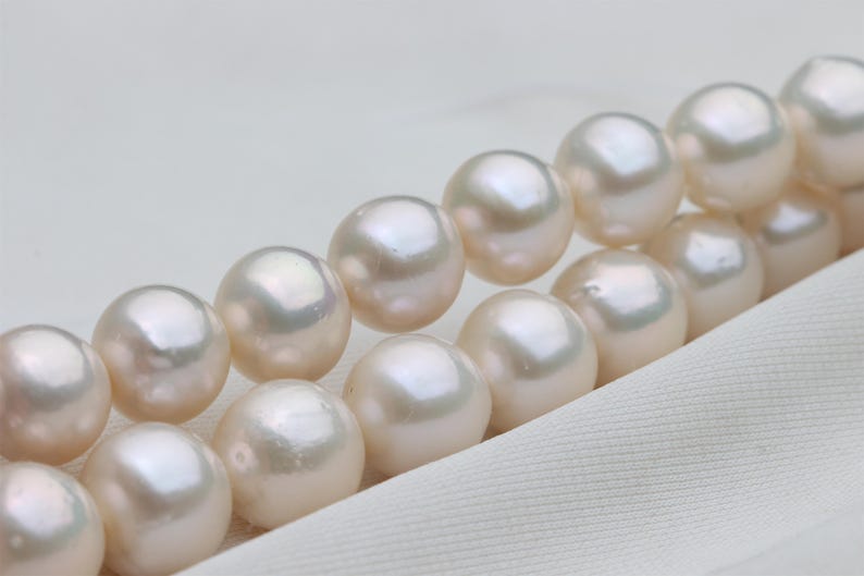 May include: Close-up of a strand of lustrous, creamy white pearls. The pearls are round and evenly sized, with a subtle iridescence. The pearls are arranged diagonally against a white fabric background, suggesting a necklace or bracelet.