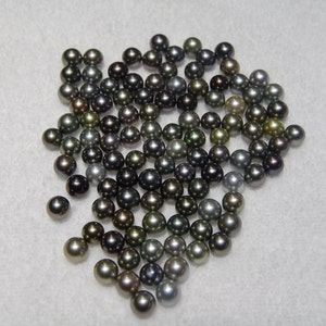 May include: A collection of black, green, and silver pearls. The pearls are round and smooth, and they are arranged in a loose pile.
