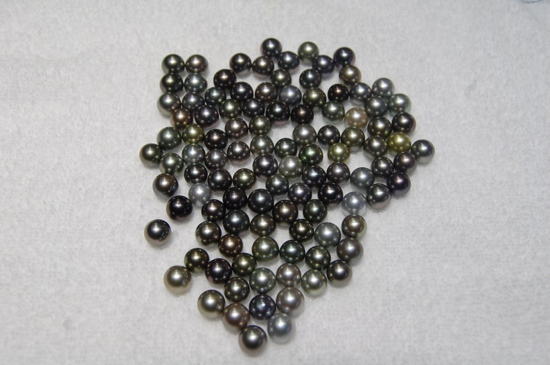 May include: A collection of black, green, and silver pearls. The pearls are round and smooth, and they are arranged in a loose pile.