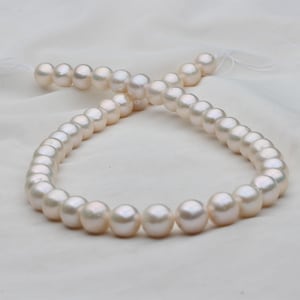 May include: A strand of lustrous, off-white pearls, each approximately 0.3 inches in diameter, lies on a white, textured fabric. The pearls are strung together, creating a necklace or bracelet. The lighting highlights their smooth, round surfaces.