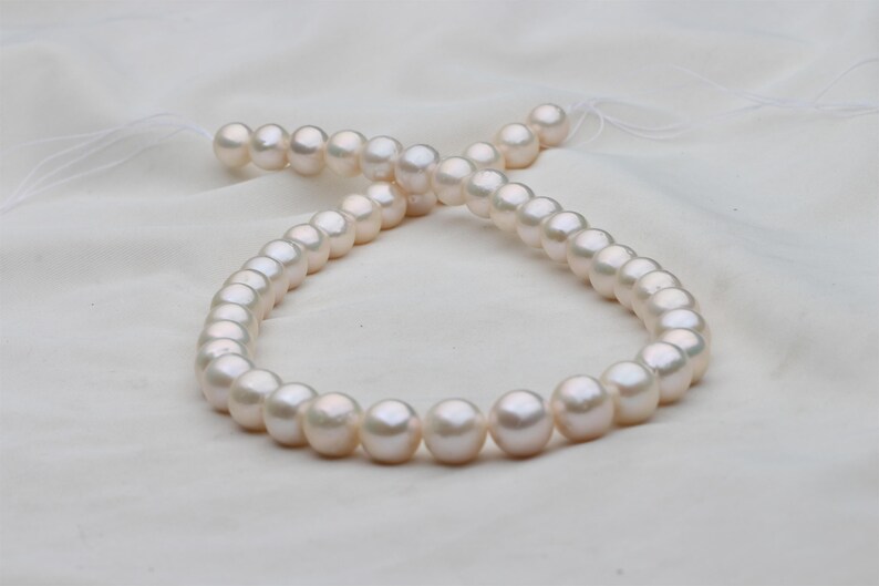 May include: A strand of lustrous, off-white pearls, each approximately 0.3 inches in diameter, lies on a white, textured fabric. The pearls are strung together, creating a necklace or bracelet. The lighting highlights their smooth, round surfaces.