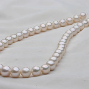 May include: A strand of lustrous, off-white pearls. The pearls are round and evenly sized, strung together on a delicate thread. The background is a soft, white fabric, highlighting the pearls' subtle sheen. A classic jewelry item.