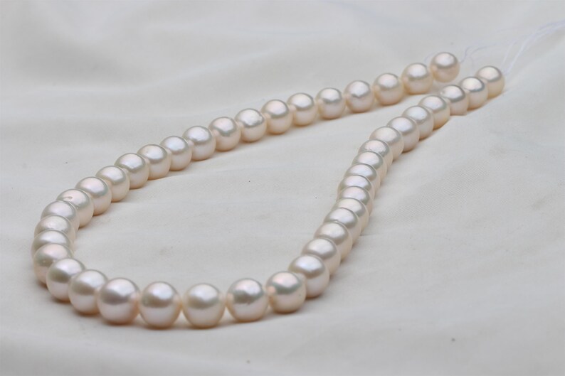 May include: A strand of lustrous, off-white pearls. The pearls are round and evenly sized, strung together on a delicate thread. The background is a soft, white fabric, highlighting the pearls' subtle sheen. A classic jewelry item.