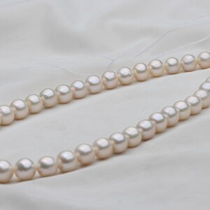 May include: A strand of lustrous, off-white pearls. The pearls are round and evenly sized, strung together on a delicate thread. The necklace is displayed on a white, textured fabric, creating a soft, elegant aesthetic.