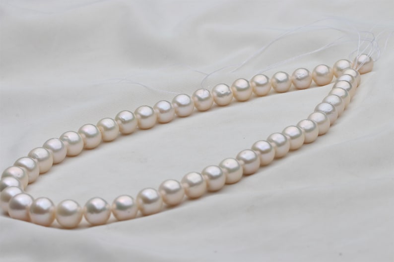 May include: A strand of lustrous, off-white pearls. The pearls are round and evenly sized, strung together on a delicate thread. The necklace is displayed on a white, textured fabric, creating a soft, elegant aesthetic.