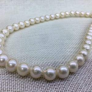 May include: A strand of lustrous, creamy white pearls. The pearls are round and evenly sized, strung together to form a necklace. The background is a light-colored, textured fabric.
