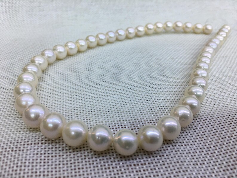 May include: A strand of lustrous, creamy white pearls. The pearls are round and evenly sized, strung together to form a necklace. The background is a light-colored, textured fabric.