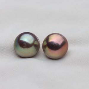 May include: Two iridescent, round pearls. One pearl has a green and silver sheen, while the other displays a pink and gold hue. The pearls are set against a white, textured background.